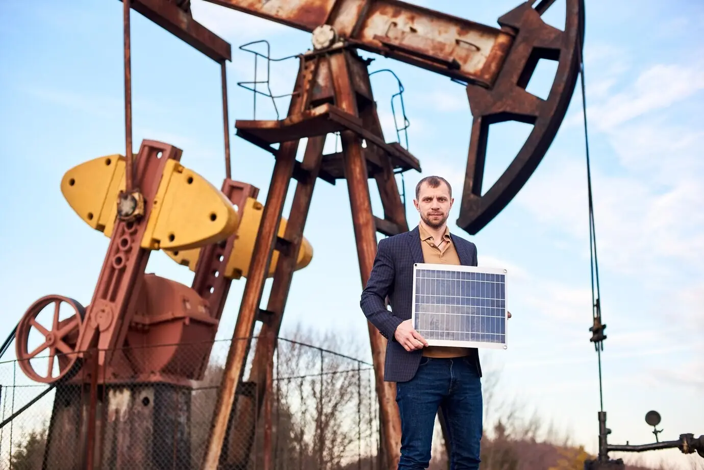 A businessman stands on an oil field, holding a mini solar module beside an oil rig.